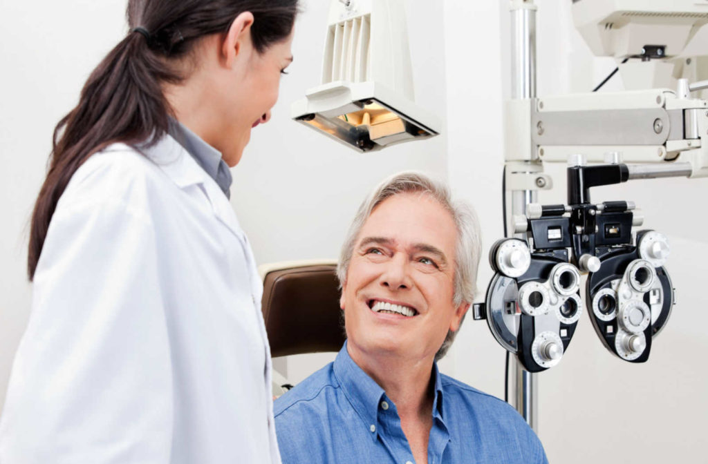 An older male patient sitting in an exam chair looks up and smiles at his female optometrist.