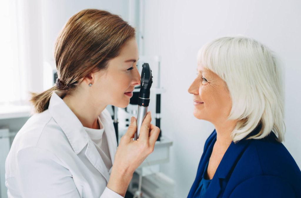 A female doctor is holding an ophthalmoscope in her right hand near her face(an apparatus used to detect and evaluate symptoms of retinal detachment or eye diseases) and examining an old patient's eye by shining the ophthalmoscope light into the patient's pupil.