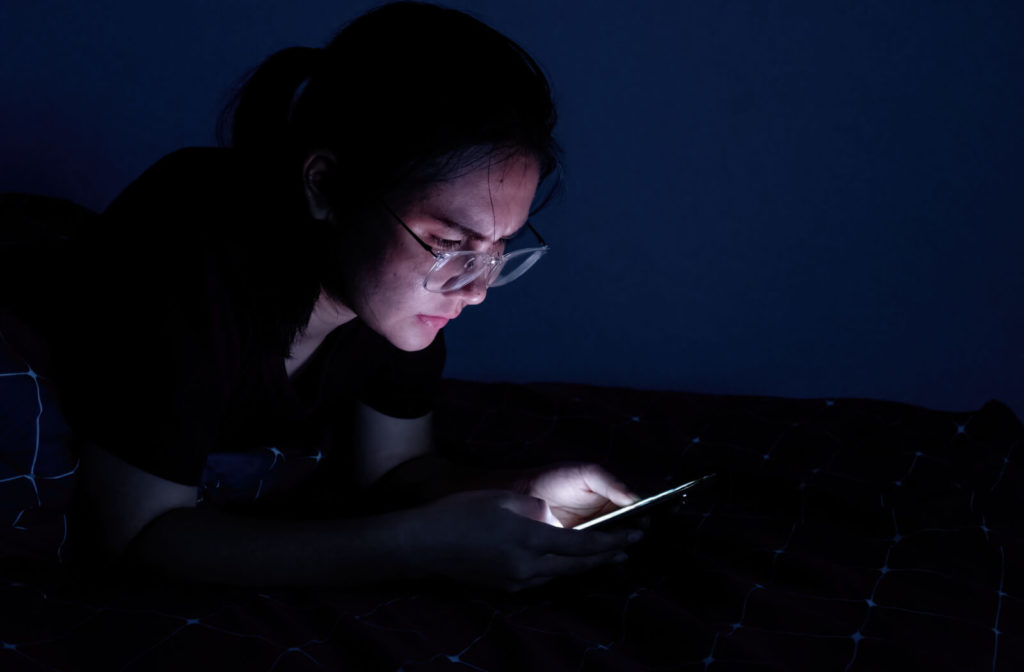 A young woman is wearing blue light glasses while using her smartphone in the dark.