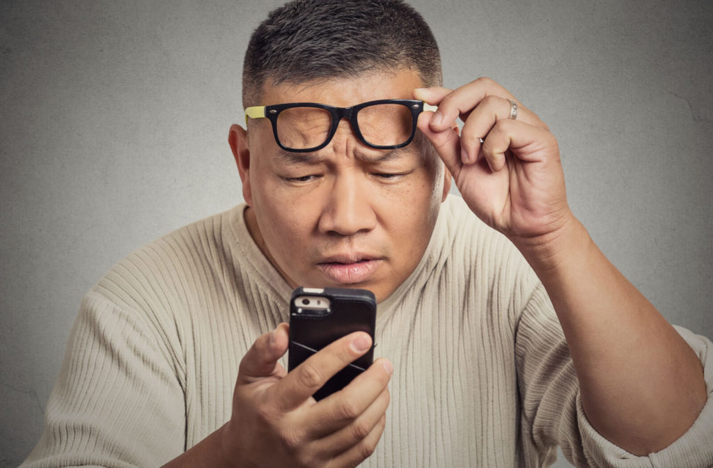 A man pushing his glasses up with his left hand and holding his smartphone very close to his face.