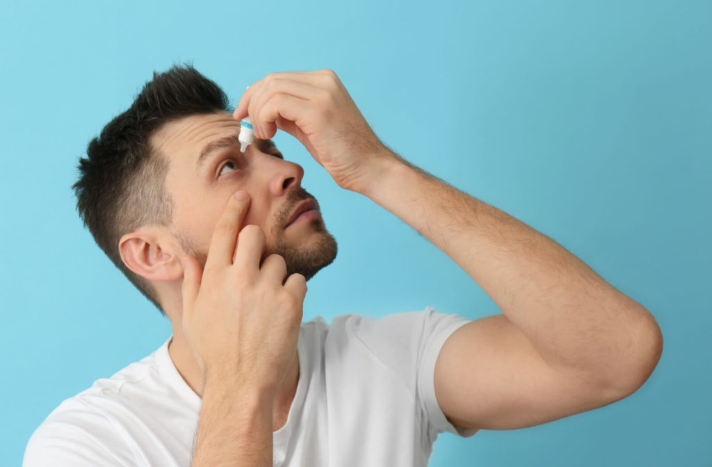A young man standing against a blue background and looking up as he applies eye drops to his right eye