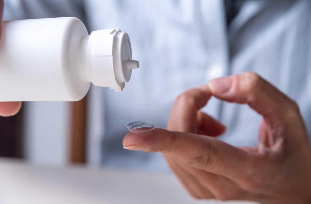 A close up of a contact lens on a fingertip and a bottle of contact solutions being poured onto it.