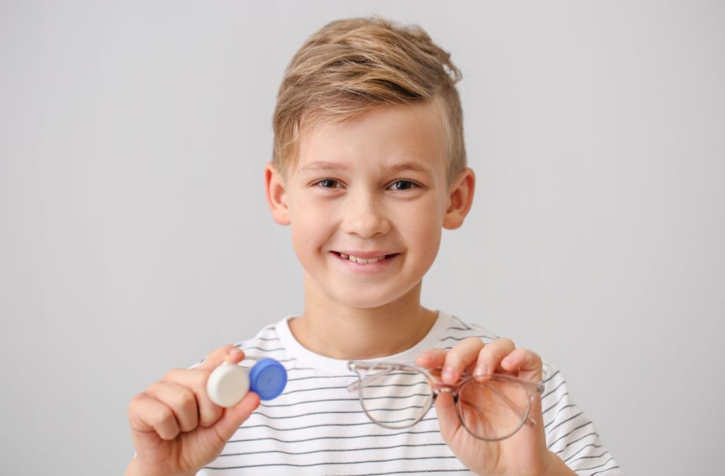 A smiling young boy holding myopia control contact lenses and eyeglasses.