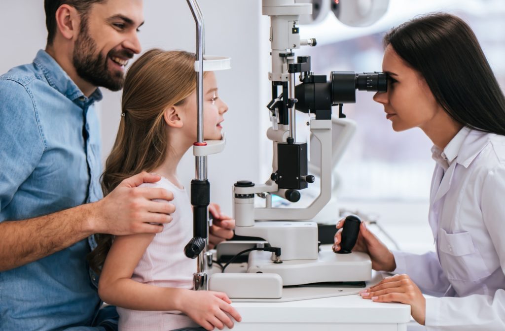 A smiling father holding his daughter on his lap while a female optometrist examines her eyes.