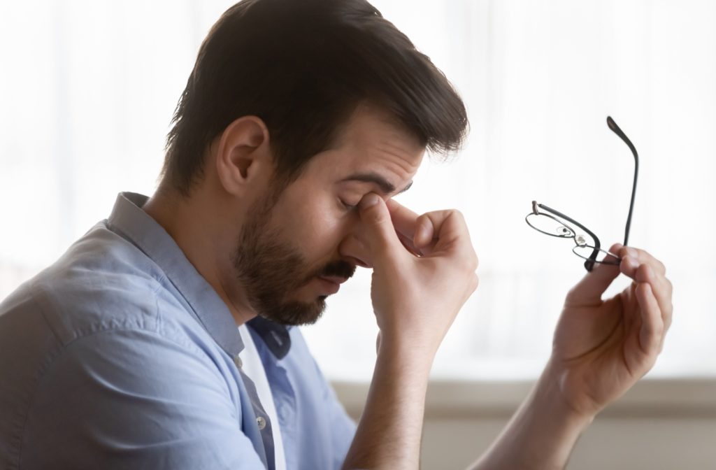 A young adult taking off their glasses to rub their eyes in frustration due to depth perception issues from astigmatism.