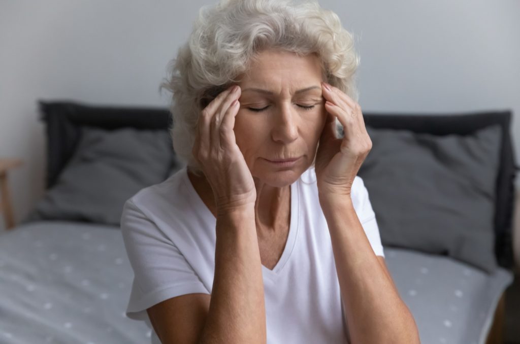 An older woman with eyes closed sitting on their bed in the morning with hands on their temples from a headache.