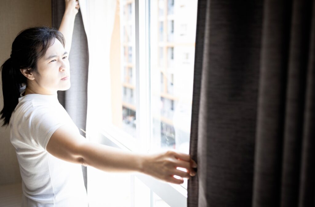 A person squints while closing heavy curtains in front of a window to block harsh, glaring light.