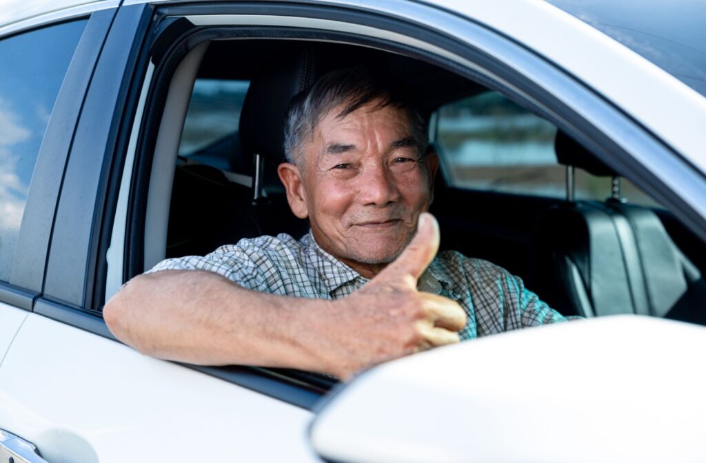 An older adult sits in the driver’s seat of a car with the window down, smiling and giving a thumbs-up.