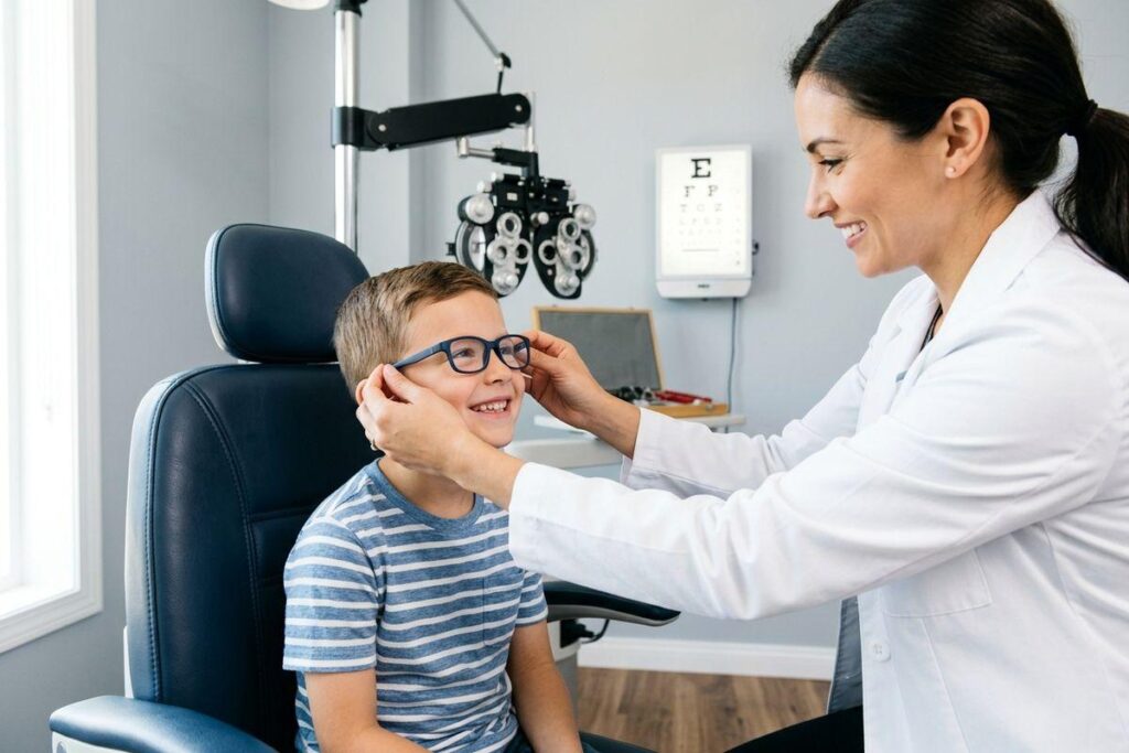 Optometrist fitting glasses on a smiling child seated in an exam chair, with eye chart and phoropter behind.